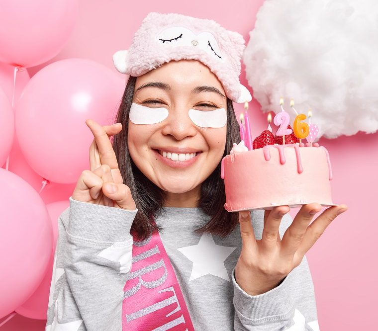 Positive Asian woman makes wish before blowing candles on birthday cake crosses fingers smiles gladfully dressed in pajama sleepmask applies beauty pads celebrates 26th bday at home balloons behind